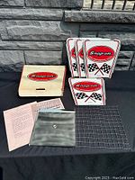 Full view of all parts included in the Snap-On vintage BBQ grill set displayed outside on black cloth against a stone wall backdrop. Visible are metal panels with Snap-On logos, grill grate, metal tray, wooden box, and printed papers.