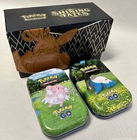 Two closed Pokémon GO tin boxes with Blissey and Snorlax designs placed next to a Pokémon Shining Fate collector box.