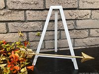 Metal decorative arrow with gold finish resting on white A-frame stand against brick background.