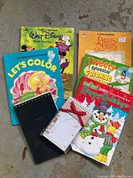 Image showing five colorful children's coloring books, a black spiral notebook, and a new notepad with a snowman festive design tied with a red ribbon.