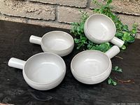 Four light gray round ceramic ovenware dishes with handles, stacked and placed on a dark surface with green plant in the background