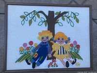 Front view of a framed needlepoint art showing a boy and girl doll holding hands under a tree with flowers.