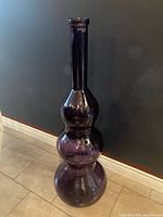 Tall purple glass floor vase with a three-tiered bulbous shape standing on a tile floor against a dark wall.