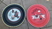 Two round metal platters side by side on floor: left black with coffee pot and cup, right red with chef grilling