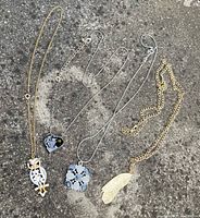 Four costume jewellery necklaces displayed on a concrete surface, showing chain variety and pendant designs including owl, heart, cluster, and feather shapes.