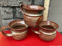 Three handmade pottery mugs set against a stone wall with a ruler for size reference, showing shape, glaze pattern and handles.