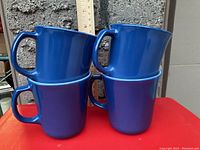 Four Pyrex mugs stacked in front of a brick wall with a ruler showing height, deep blue color with handle and white interior visible.