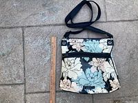 Full view of the handbag placed on a stone surface alongside a ruler showing size, floral pattern with blue, pink and white flowers on black background, black shoulder strap.