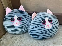 Two round gray and black striped plush cat stuffed animals with pink ears shown side by side against a textured wall backdrop.