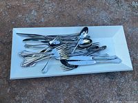 Photo of a white rectangular tray filled with various silver-toned cutlery including knives, forks, spoons, and a gravy sauce spoon on a concrete surface.