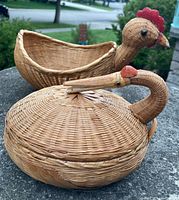 Vintage wicker baskets shaped like a chicken and a goose placed outside on a surface, showing the chicken basket with open top and the goose basket with removable lid and locking peg.