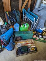Photo showing small frame pack, two blue camp chairs, blue sleeping pad, green 5-gallon water jug, green metal toolbox, insulated bags, barbecue tools, and other camping gear items.