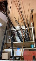 Multiple fishing rods standing upright, tightly placed together on a shelf with other fishing equipment below.