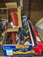 Wide view of two boxes showing various hand tools including handles of screwdrivers, pliers, a red-handled manual drill, and a cardboard box with a red plastic case for the screwdriver set inside.