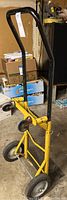 Front-right angled view of black and yellow hand truck showing curved black handle, yellow frame, rear wheels and front casters, mesh panel and footplate.