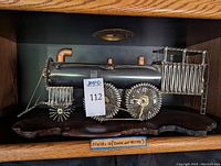 Side view of metal train-shaped clock showing cylindrical black body and coil wire wheels mounted on the wooden base