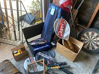 Various sports equipment laid out near a gate and on concrete floor including badminton kit bag, racquetball racket, flags in carrying cases, hockey sticks, dart board, and other items.
