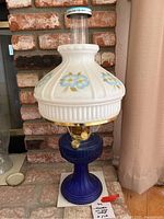 Full view of the Aladdin oil lamp with blue ribbed base and white milk glass shade with blue and yellow flowers, in front of brick background.