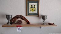 Wide view of shelf mounted on wall displaying two metal goblets, two boomerangs with Aboriginal designs, small clay bowl, and framed art print above.