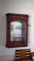 Front view of the vintage wooden wall display cabinet mounted on the wall, showing glass door, brass latch, and interior glass shelving.