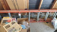 Photo showing orange extension ladder with metal rungs against a wall with some boxes around.