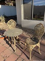 Outdoor image showing one round wrought iron bistro table and two matching wrought iron chairs on a tiled patio area in natural light.