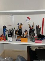 Shelf showing various office supplies including pens, pencils stored in Italian and marble vases, tape rolls, envelopes, and small office tools.