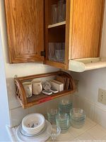 Kitchen corner countertop with stacked Pyrex custard bowls, white dishes, mugs, thermometers in a rattan shelf, and glass drinking cups inside wooden cabinet.