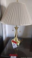 Full view of Stiffel brass lamp with beige pleated fabric shade and pull chains, sitting on glass table.