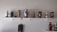 Full view of all 9 souvenir vases displayed on white wall shelf fixed with brackets.