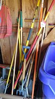 Multiple yard tools including pitchfork, shovel, and other long handled tools leaning against wooden wall in shed.