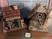 Photo showing two handmade wooden bird houses side by side. Left one has shingled roof, right one has bark roof with attached branches and signs.