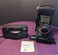Photo shows the Kodak No1 Diomatic folding camera with bellows extended beside the Minolta Freedom AF 35 black compact camera on purple surface.