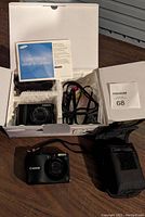 Samsung NV100 HD camera inside original box with user manual, cables, and accessories, Canon Powershot A1200 with camera case beside it.
