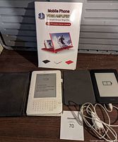 Photo showing both white Kindle e-readers in black and gray cases, the USB charging cord, MaxSold tag, and boxed red mobile phone screen magnifier.