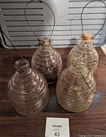 Four large ribbed glass wasp catchers in varying condition shown on wooden surface with gray background. Two have wire handles and cork tops, two have no tops with exposed openings. Some dirt visible on two catchers.