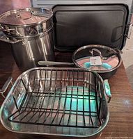 Cookware set displayed on table including roasting pan with rack, stock pot with lid and strainer, frying pan with lid, and griddle.