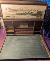 Open folding antique wooden desk showing black slate writing surface and multiple storage sections, with printed text logo on upper panel.
