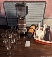 Full view of the lot items on a table including blender, six champagne flutes, drawer protector mat roll, potato ricer, and Tupperware container with orange lid