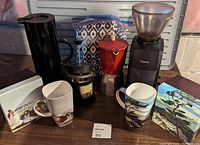Overview of the lot showing four mugs with Group of Seven landscape designs, black carafe, French press, stovetop espresso maker, coffee grinder, and folded patterned tablecloth in the background.