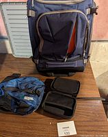 Two-in-one convertible rolling backpack shown standing upright; blue Mountain Co-Op cycle fanny pack and black electronic case displayed in front on table.