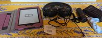 Kindle e-reader powered off showing low battery symbol, pink textured fabric case unfolded, Sony wired black over-ear headphones with coiled cord, and Outman monocular with carrying case and cleaning cloth.