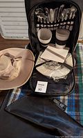 Picnic backpack open showing cutlery sets, plastic cups, plates, and netted storage inside. Beige safari hat beside backpack. Blue and brown plaid travel mat under backpack with black carrying case.
