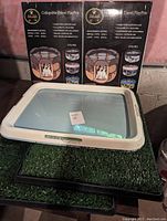 Neat'n Dry pet pad holder on top of one artificial grass mat with two boxed collapsible travel playpens behind.