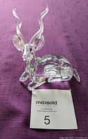 Front-view photo of the clear Swarovski Kudu figurine sitting on a purple fabric background, showing curved crystal horns and faceted body detailing.