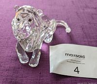 Front view of clear Swarovski crystal lion figurine on purple fabric, displayed alongside a MaxSold identifier label with number 4.