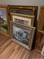 View showing multiple framed artworks stacked against a wall on the floor, featuring ornate frames and a range of sizes.