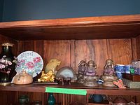 Full shelf view showing cloisonne vase, porcelain plate, ceramic trinket box, carved stone figurines, Laughing Buddha, agate slice, monk figurines, teacups