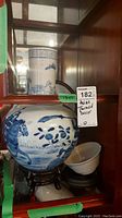Shelf display of large blue and white porcelain baluster vase on carved wood stand, smaller cylindrical brush pot behind, and white bowl below