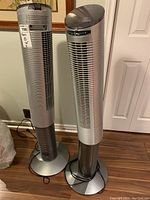 Two matching silver Windchest tower fans standing side by side on a wooden floor near a white wall and door.